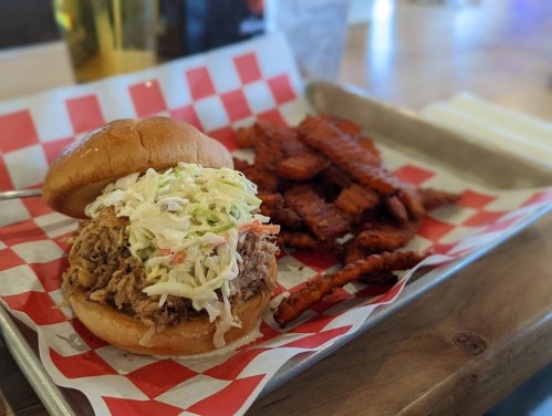 A pulled pork sandwich topped with coleslaw, served with sweet potato fries on a tray with red and white checkered paper.