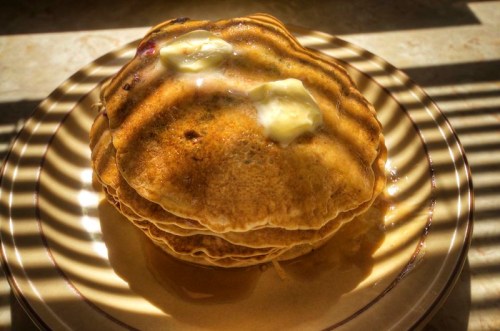 Stack of golden pancakes topped with butter, drizzled with syrup, on a plate with sunlight casting striped shadows.