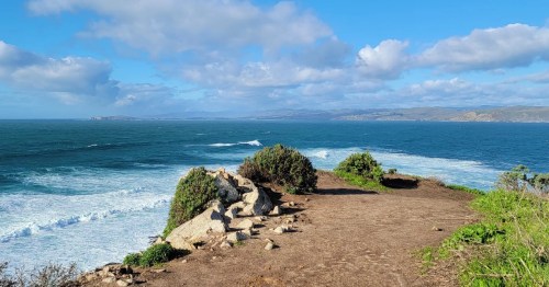 A scenic coastal view with waves crashing against rocks under a blue sky with scattered clouds.