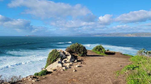 A scenic coastal view with waves crashing against rocks and greenery under a partly cloudy sky.