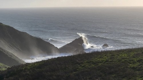 Waves crash against rocky cliffs along a serene coastline, with mist rising in the sunlight.