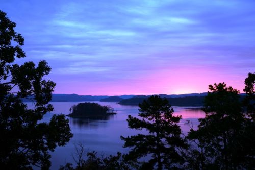 A serene lake at sunset, with vibrant purple and pink hues reflecting on the water and silhouetted trees in the foreground.