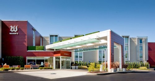 Modern hotel exterior with a glass entrance, greenery, and a clear blue sky. Welcoming atmosphere.
