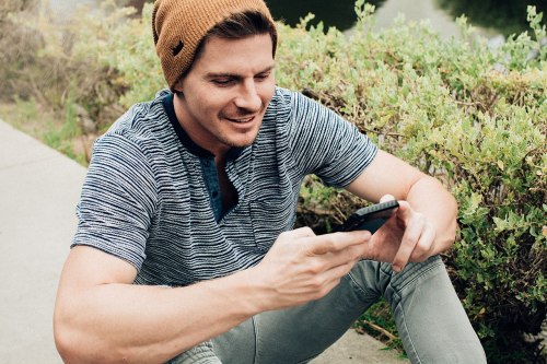 A young man in a beanie sits outdoors, smiling while looking at his smartphone. Greenery is in the background.