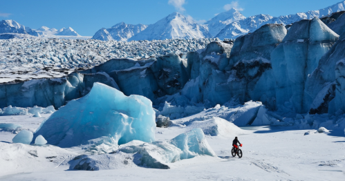 A cyclist rides through a stunning icy landscape with towering blue glaciers and snow-capped mountains in the background.
