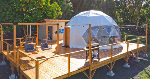 A geodesic dome on a wooden deck surrounded by greenery, featuring hanging lights and outdoor seating.