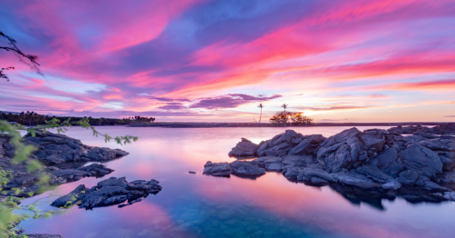 A serene landscape featuring a colorful sunset over calm waters, with rocky shores and silhouettes of trees.