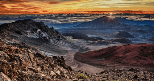 A stunning sunset over a volcanic landscape, with rugged mountains and clouds in the foreground and background.