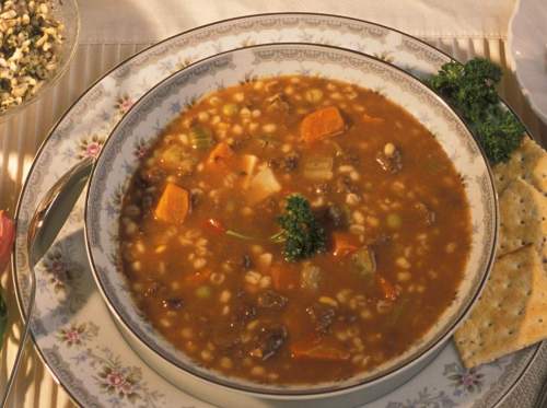 A bowl of hearty vegetable and beef soup with barley, garnished with parsley, served with crackers on a decorative plate.