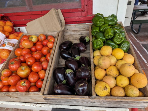 A wooden display of fresh produce featuring red tomatoes, green bell peppers, eggplants, and oranges.