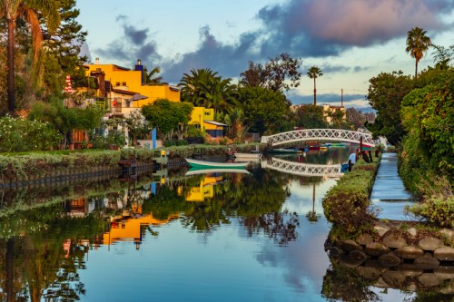 A serene canal scene with colorful houses, palm trees, and a white bridge reflecting in the water at sunset.