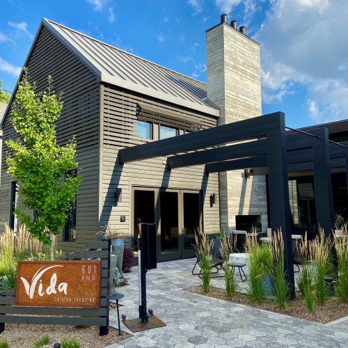 A modern restaurant exterior with a wooden facade, large windows, and a sign reading "Vida" surrounded by greenery.