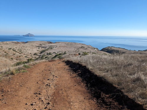 A dirt path leads through dry grassland, overlooking a calm ocean and distant island under a clear blue sky.