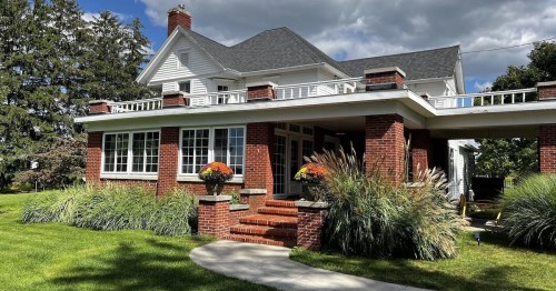 A charming brick house with a porch, surrounded by greenery and colorful flowers under a partly cloudy sky.