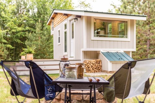 A cozy tiny house in a forest setting, with outdoor seating and a table in front, surrounded by trees.