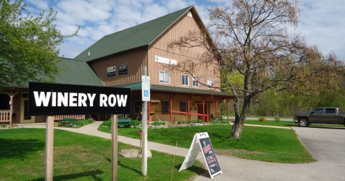 A rustic winery building with a sign reading "Winery Row" and a grassy area in front, under a partly cloudy sky.