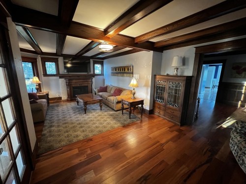 Cozy living room with wooden beams, a fireplace, a sofa, and a coffee table on hardwood floors. Natural light from windows.