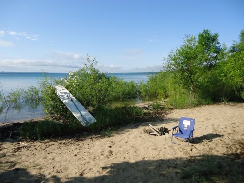 A sandy beach with a blue chair, a fire pit, and lush greenery by the water under a clear sky.