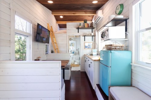 Cozy tiny home interior featuring a kitchen, dining area, and loft bed with wooden accents and a retro blue fridge.