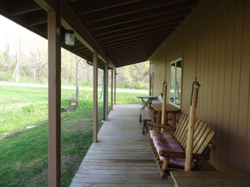 A wooden porch with swings and a table, surrounded by green grass and trees in the background.