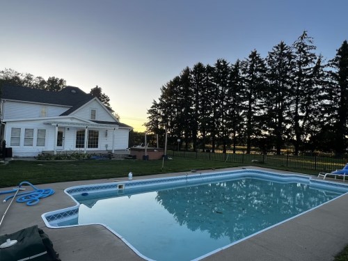 A serene backyard scene featuring a swimming pool, a white house, and tall trees at sunset.