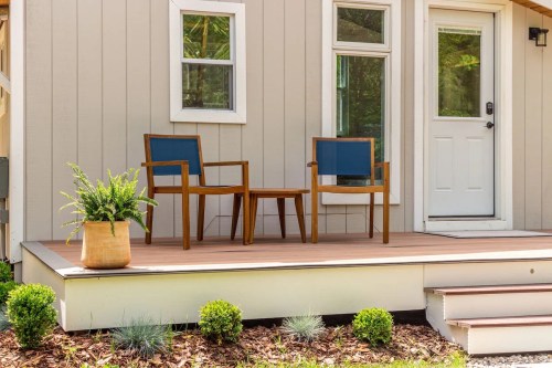 A cozy porch with two wooden chairs, a small table, and a potted plant, surrounded by greenery.