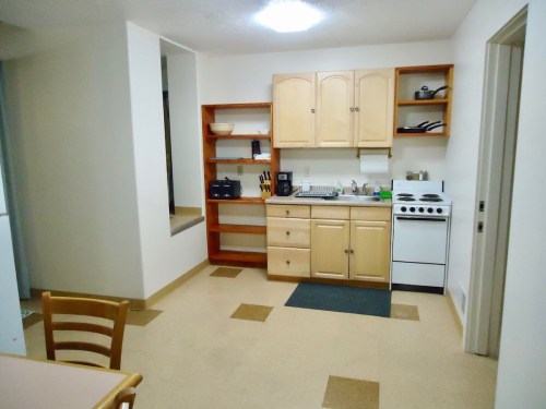 A simple kitchen with wooden cabinets, a stove, sink, and a small dining table in a light-colored room.