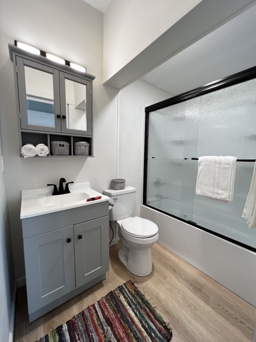 A modern bathroom featuring a gray vanity, toilet, shower with frosted glass, and a colorful rug on the floor.