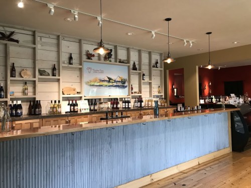 A rustic wine bar with a wooden counter, shelves of bottles, and artwork on the wall, featuring warm lighting.
