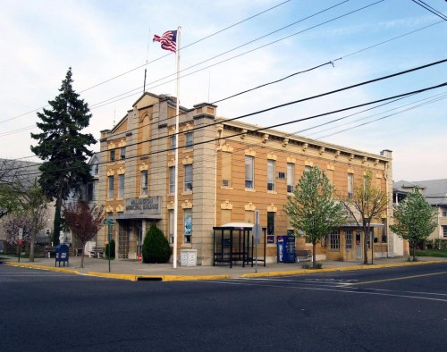 Historic building with a flag, surrounded by trees and a bus stop, located at a street corner.