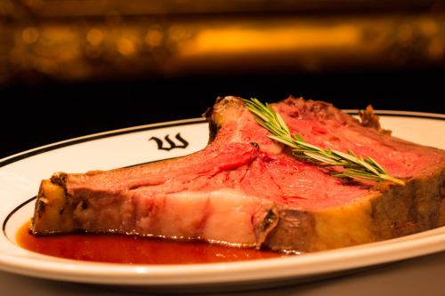 A beautifully cooked slice of prime rib on a white plate, garnished with a sprig of rosemary and served with sauce.