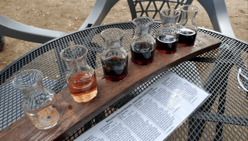 A wooden flight board with six small glass bottles of different colored liquids, placed on a metal table.