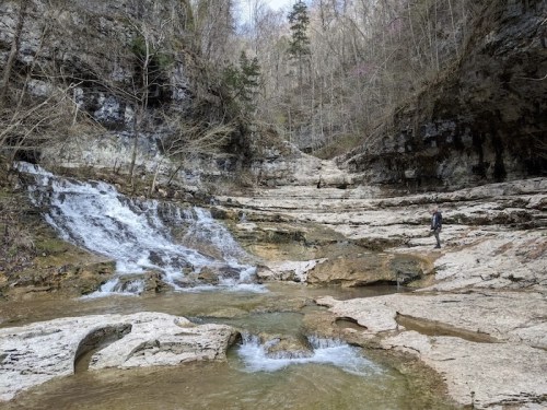 A serene landscape featuring a small waterfall and rocky terrain, with a person walking along the water's edge.