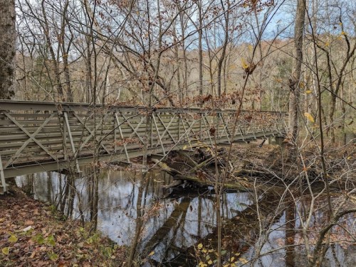 A wooden bridge crosses a calm stream, surrounded by trees with autumn leaves.
