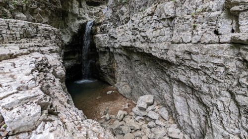 A serene waterfall cascades into a rocky pool, surrounded by steep, textured stone walls in a natural canyon.