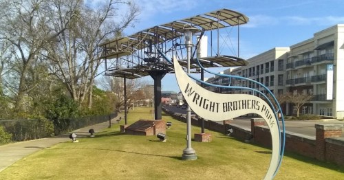 Wright Brothers Park featuring a large sculpture of an airplane and a sign, with green grass and buildings in the background.