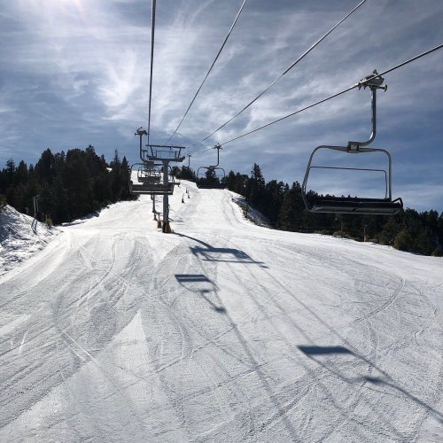 A ski lift ascends a snowy slope surrounded by trees under a clear blue sky.