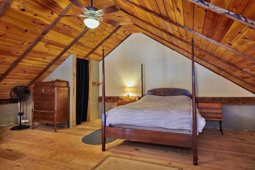 Cozy attic bedroom with wooden beams, a four-poster bed, dresser, and a fan, featuring warm, rustic decor.