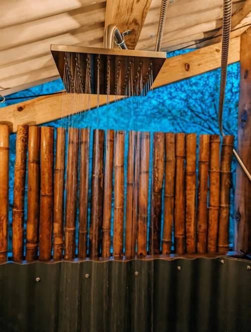 A modern showerhead with water cascading down, set against a bamboo wall and a twilight sky.