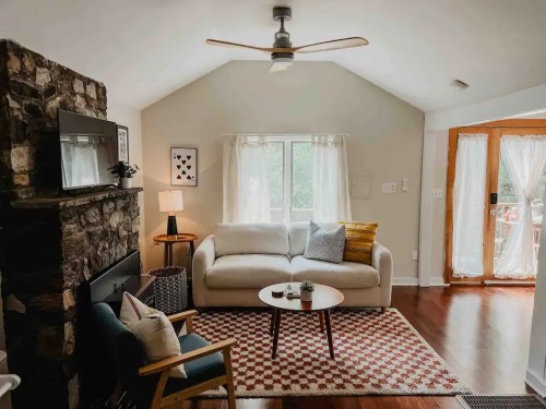 Cozy living room with a stone fireplace, white sofa, wooden coffee table, and large windows letting in natural light.