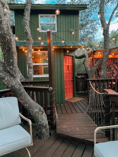 A cozy treehouse with a bright red door, surrounded by trees and string lights, on a wooden deck.