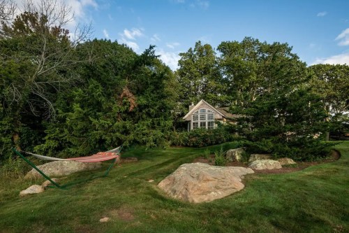 A cozy house surrounded by trees and rocks, with a hammock set up in a lush green yard under a blue sky.