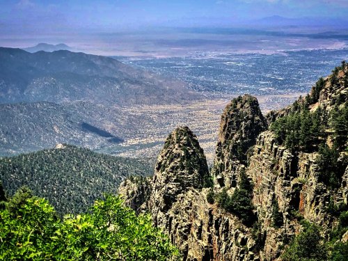A panoramic view of rugged mountains and valleys under a clear blue sky, with lush greenery in the foreground.