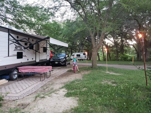 A child rides a pink bike near a camper and parked car, surrounded by trees and a grassy area at dusk.