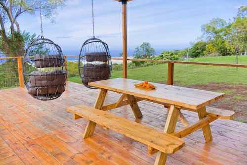 A wooden deck with a picnic table and two hanging chairs, overlooking a scenic landscape and ocean view.