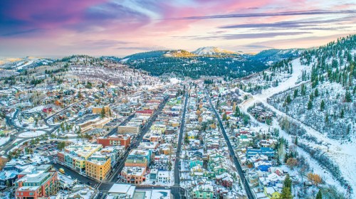 Aerial view of a snowy town surrounded by mountains, with colorful buildings and a vibrant sunset sky.