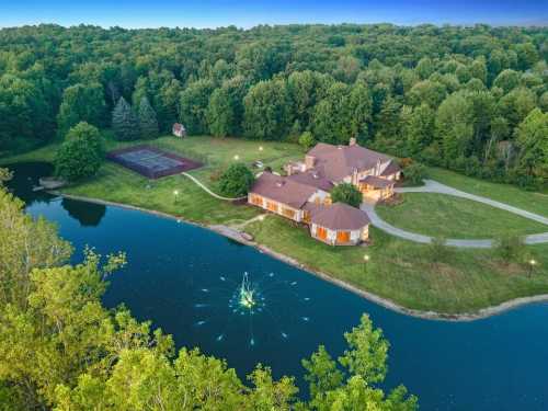 Aerial view of a spacious home by a pond, surrounded by trees, with a tennis court nearby and lights reflecting on the water.