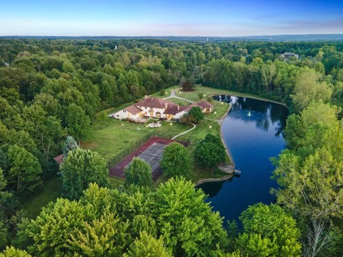 Aerial view of a spacious home surrounded by lush greenery and a serene pond, with a tennis court nearby.