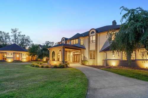 A spacious, elegant home with a curved driveway, surrounded by lush greenery and soft evening lighting.