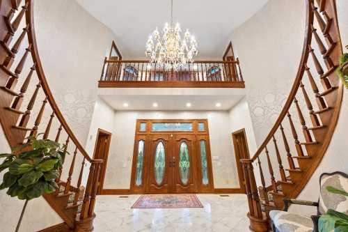 Elegant foyer with a grand chandelier, curved staircases, and double doors, featuring marble flooring and decorative plants.